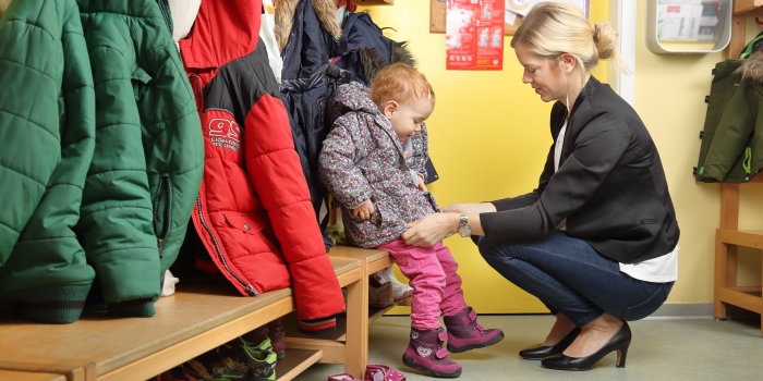 Foto: Abholen aus der Kita © RioPatuca Images/Fotolia Eine junge Frau im Businessoutfit holt ein kleines Mädchen aus der Kindertagesstätte ab und zieht ihr die Jacke an.